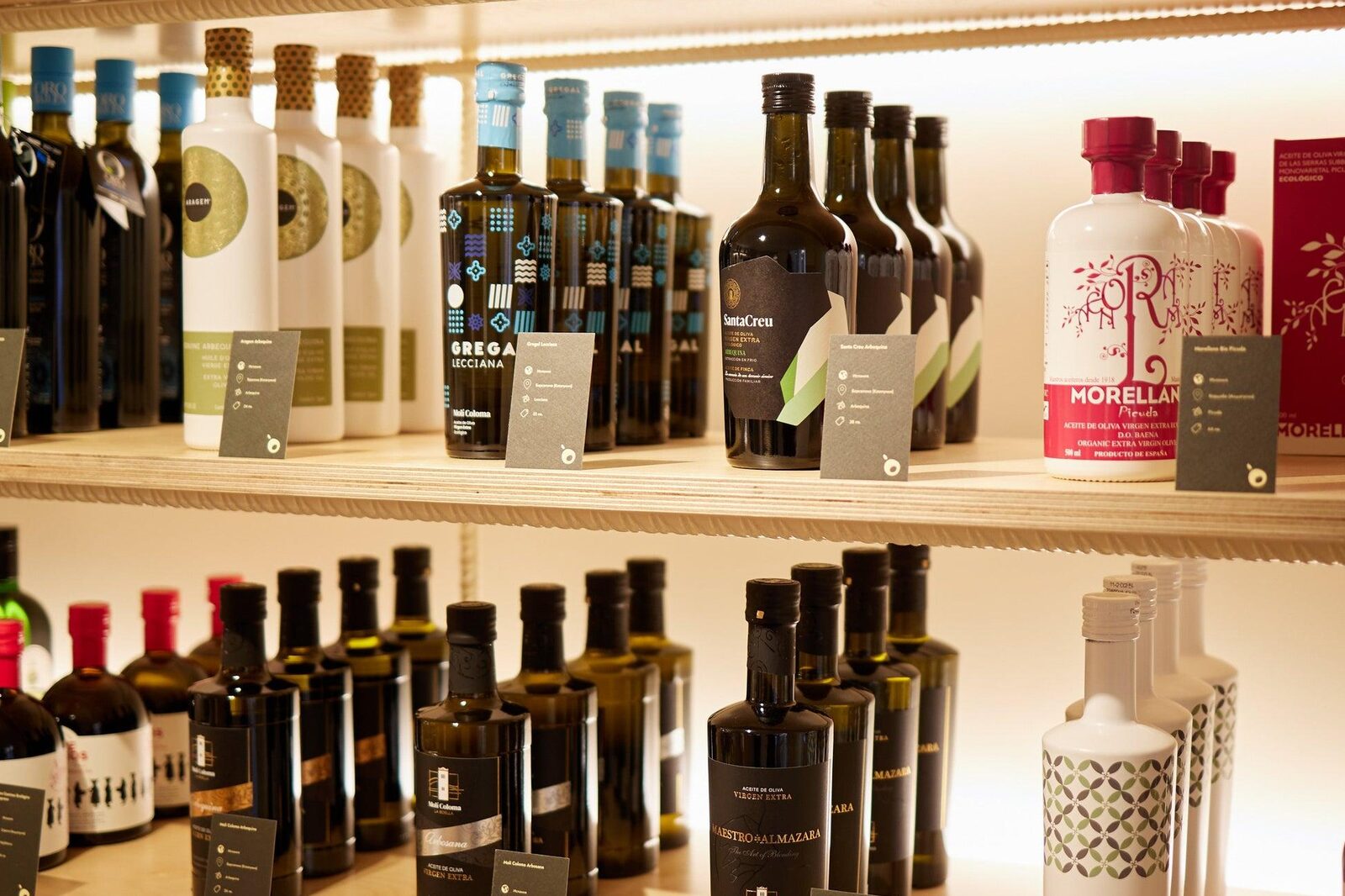 Backlit wooden shelves of Spanish extra virgin olive oils in a specialist shop, Gregal Lecciana, Santa Creu, Morellana Picuda and Maestro de Almazara among them, each with a small grey card naming the oil, its origin and its variety