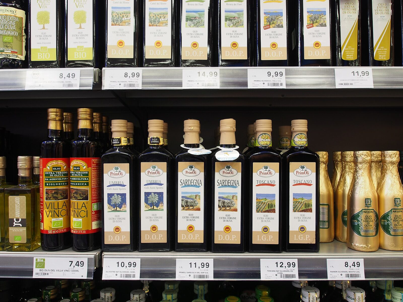 A German supermarket shelf of Italian extra virgin olive oils: PrimOli bottles labelled Umbria, Sardegna and Toscano with D.O.P. and I.G.P. seals, organic Villa Vinci bottles marked 100% Italiano and gold-wrapped bottles, with more D.O.P. oils on the shelf above and price tags from 7.49€ to 14.99€